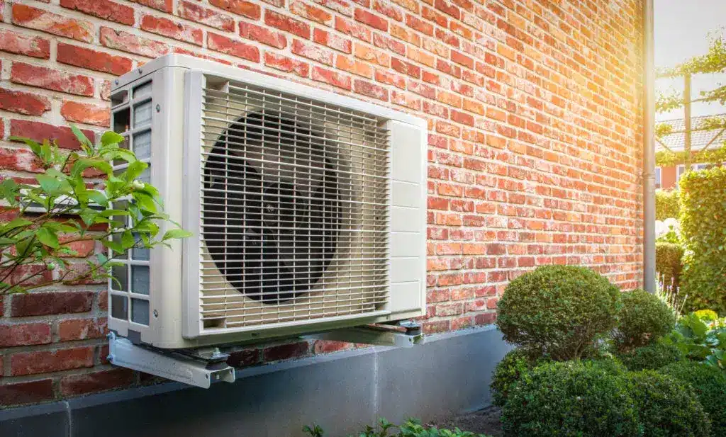 Side view of outdoor energy unit hanging on brick wall of house on a sunny day. Air to air heat pump for cooling or heating the home. Outdoor unit powered by renewable energy. Air conditioner and air heat pump
